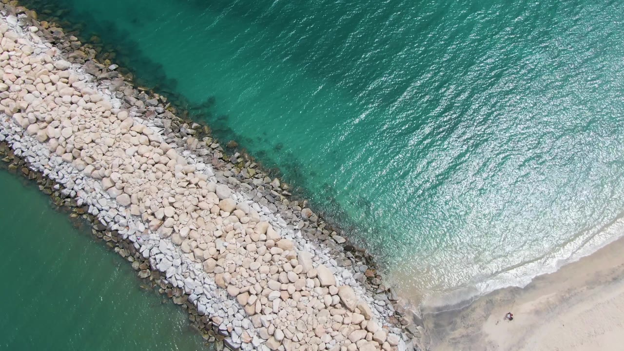 Top-down Puerto Los Cabos Beach frame with bright reflected light across rippled turquoise water, the breakwater along the left side, sand at the lower right, and no visible people.