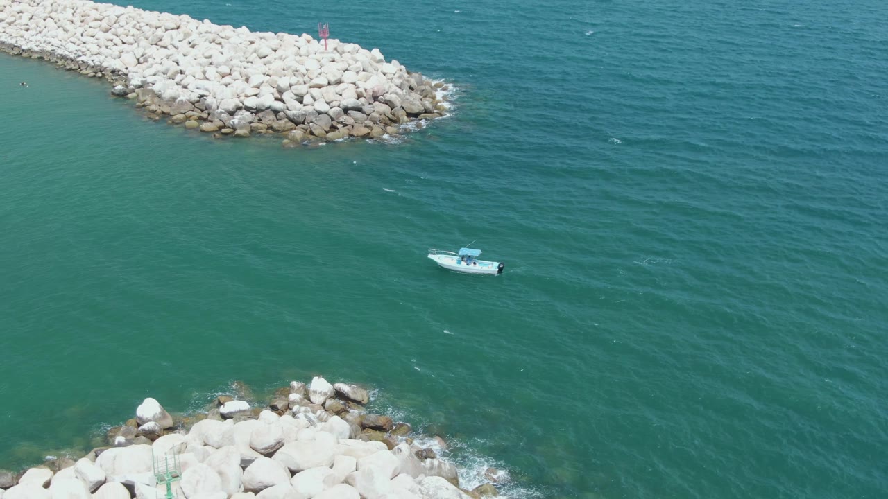 Aerial frame from Puerto Los Cabos Beach with the small boat slightly farther across the open water, pale breakwater rocks above and below, and no visible people.