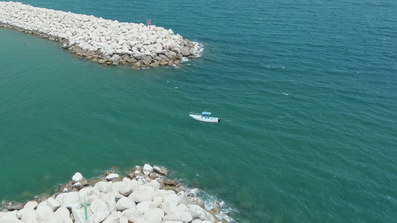 Puerto Los Cabos Beach aerial view with a white boat crossing calm green water, a long rocky breakwater at the top, foreground rocks at the bottom, and no visible people.