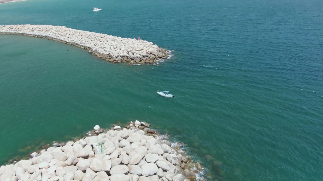 Aerial frame over Puerto Los Cabos Beach showing a curved pale rock breakwater, a second rock section at the bottom edge, green turquoise water, a small boat offshore, and no visible people.