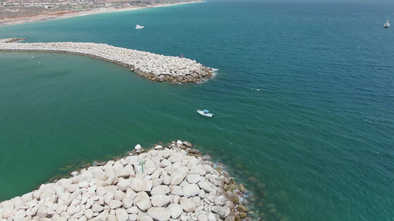 Puerto Los Cabos Beach aerial frame showing the lower breakwater occupying the foreground, the long pale rock arm across the upper water, a small boat between them, distant shoreline, and no visible people.