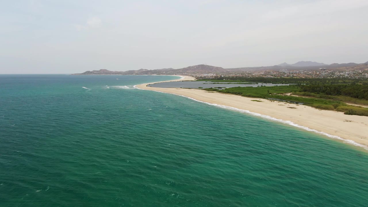 Puerto Los Cabos Beach aerial view with turquoise water in the foreground, pale sand curving along the right side, green vegetation behind the shore, distant hills, and no visible people.