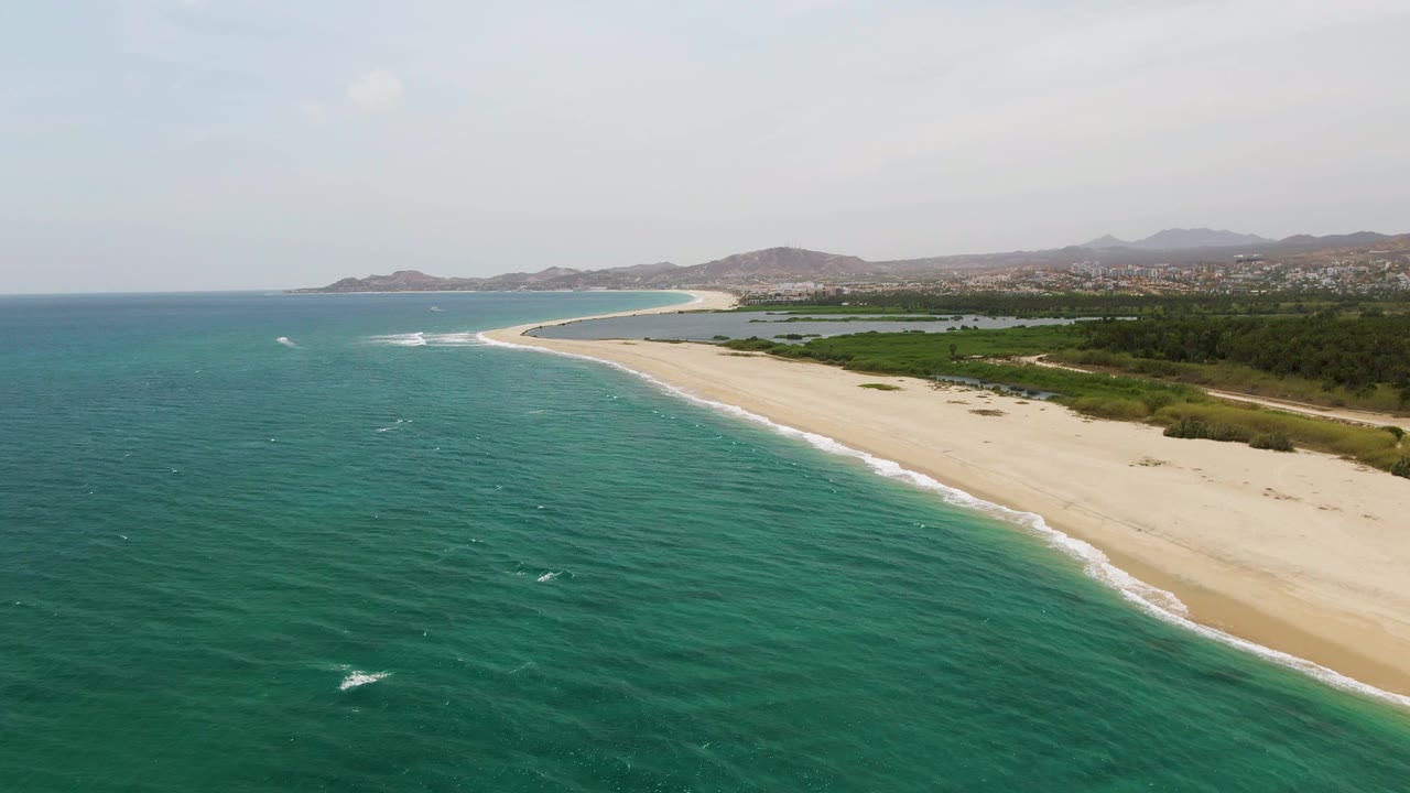 A wide aerial frame of Puerto Los Cabos Beach shows the ocean filling the left foreground, the pale beach running diagonally at right, green shoreline vegetation, distant low hills, hazy sky, and no visible people.