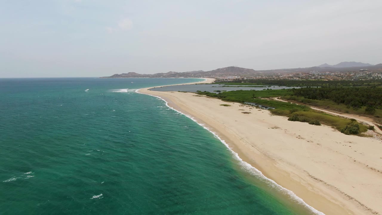 Aerial frame over Puerto Los Cabos Beach with turquoise ocean on the left, pale sand curving toward distant hills, inland greenery, and no visible people.