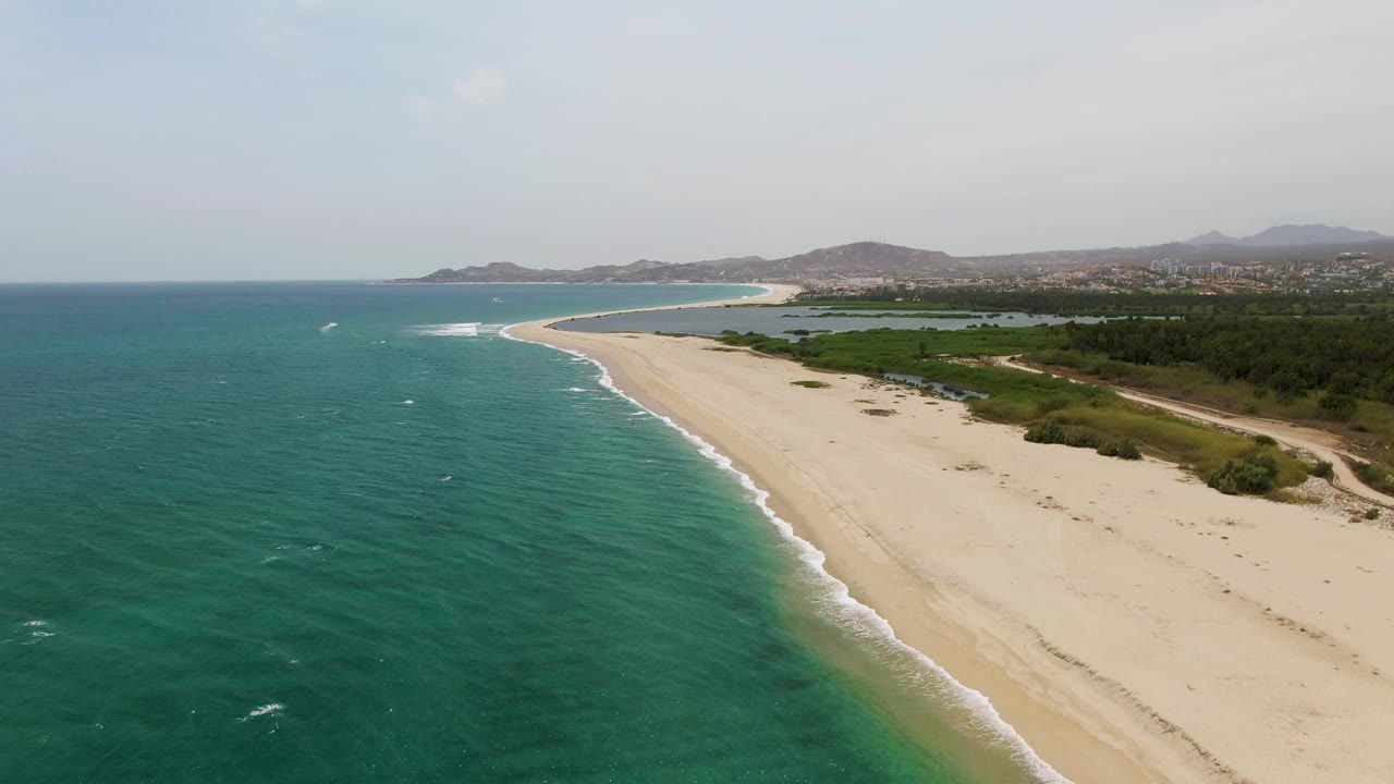 Aerial view at Puerto Los Cabos Beach with the surf line slightly farther along the diagonal sandbar, teal ocean texture, inland greenery, distant hills, and no visible people.