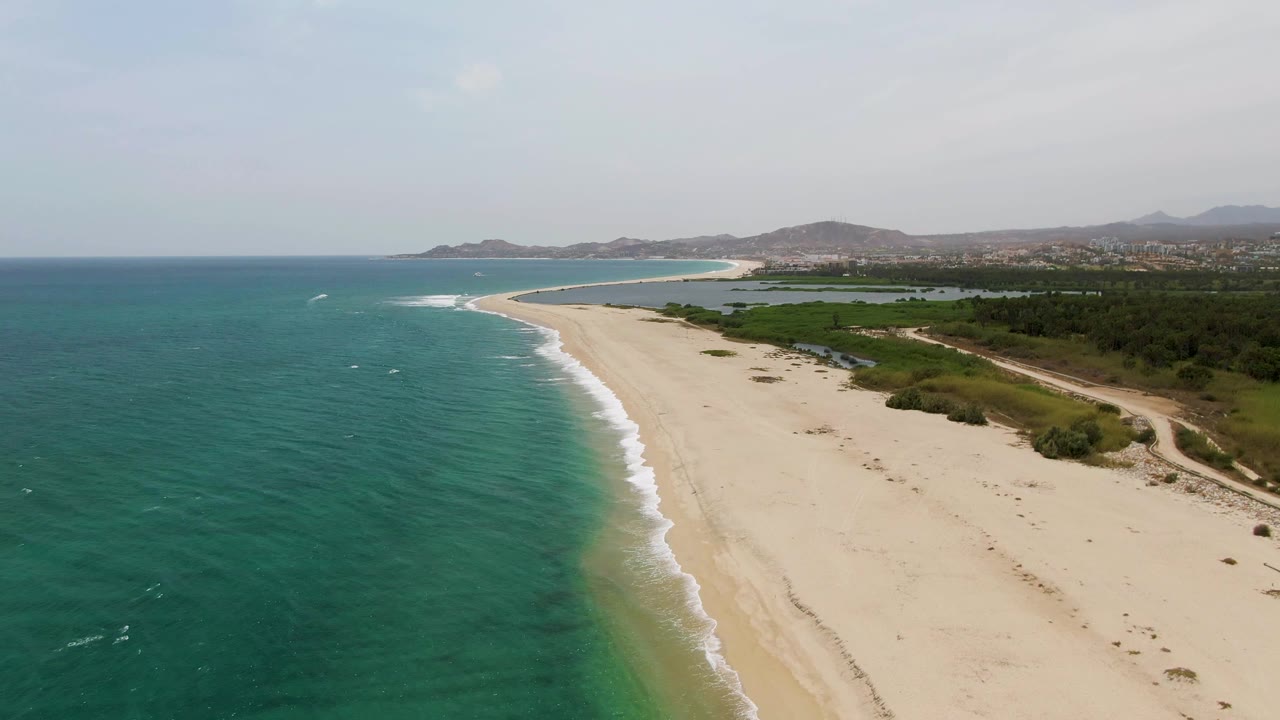 Curving sandy shoreline with ocean, surf, inland greenery, and hazy hills.