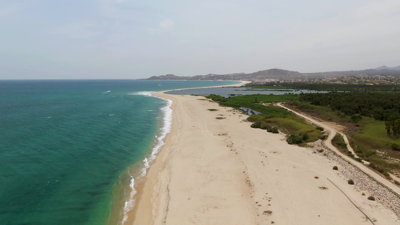 Aerial frame over Puerto Los Cabos Beach, Mexico, with turquoise ocean at left, pale sand down the center, green vegetation and a dirt path at right, distant hills, and no visible people.