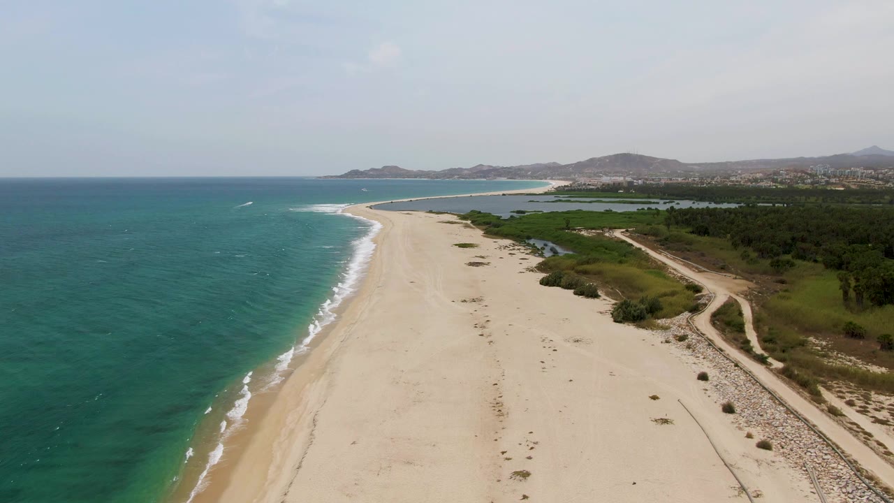 Puerto Los Cabos Beach aerial view one second in, showing the surf line edging the broad sand while the dirt road and green strip run along the right side with no visible people.