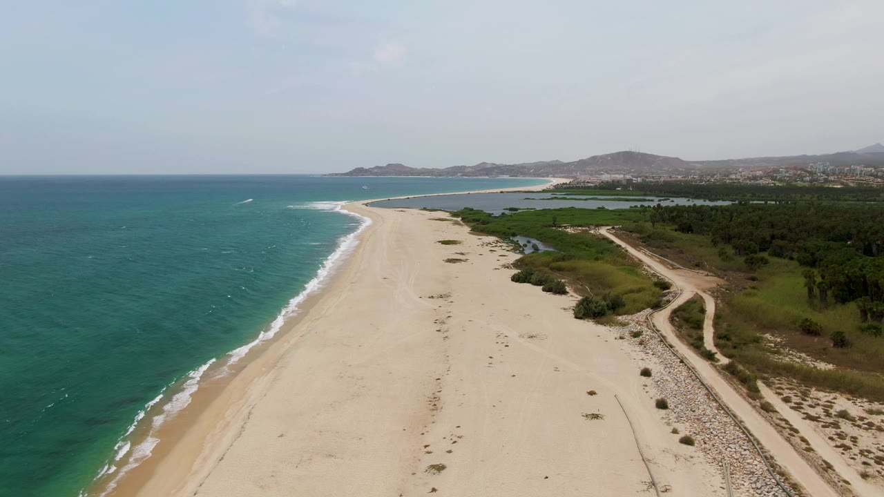 Aerial view over Puerto Los Cabos Beach with more of the curving shoreline visible ahead, turquoise water to the left, sandy beach below, greenery to the right, and no visible people.