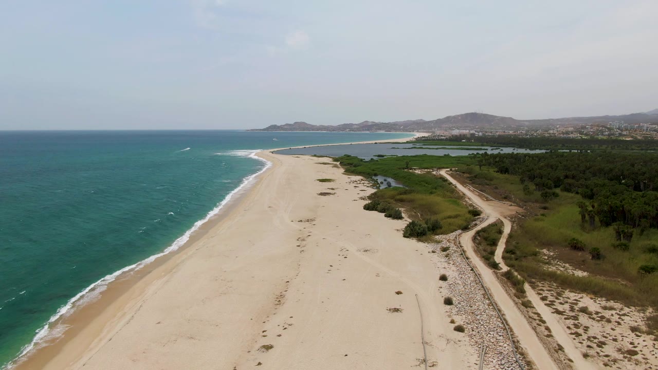 Aerial coastline view with empty sand, ocean waves, dirt path, and vegetation.