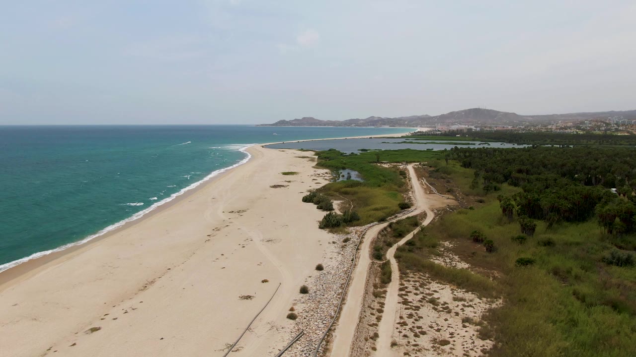 Aerial view over Puerto Los Cabos Beach with turquoise ocean on the left, pale sand, a dirt coastal road beside green vegetation, distant hills, and no visible people.