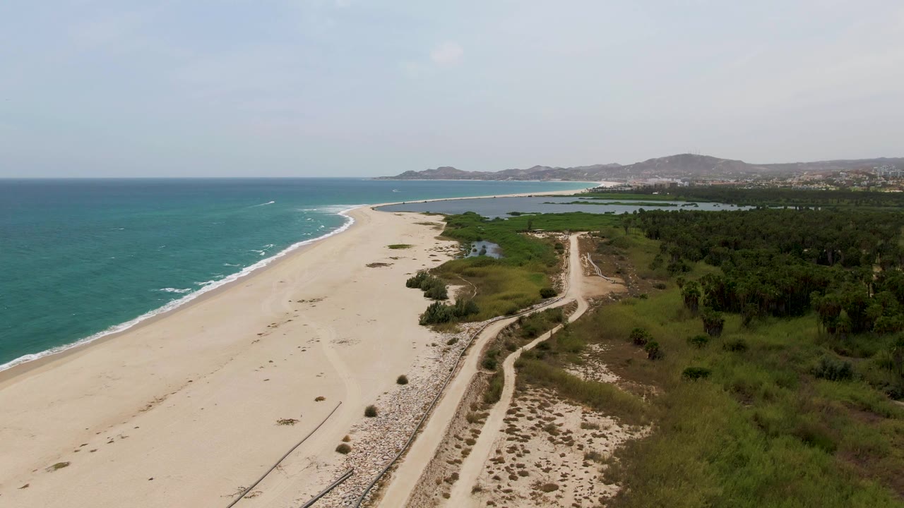 High aerial frame at Puerto Los Cabos Beach showing the shoreline curving ahead, surf along the left edge, road tracks through sand and greenery, distant terrain, and no visible people.