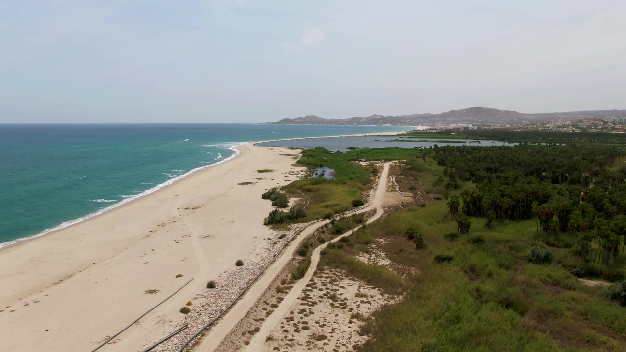 Aerial frame from Puerto Los Cabos Beach with turquoise water, light surf, broad sand, a road through the right side, dense green vegetation, distant coastal development, and no visible people.