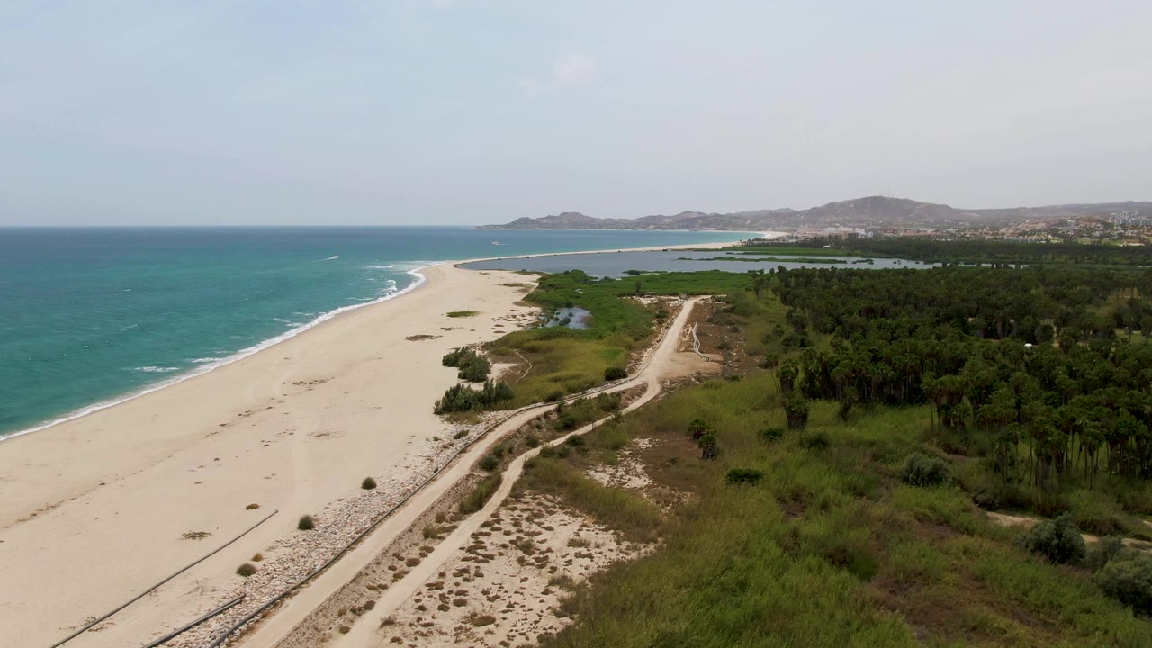 Drone perspective over beach, shoreline surf, dirt road, vegetation, and hazy hills.