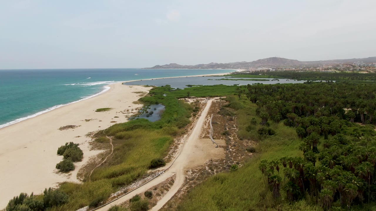 Aerial view of sandy beach, turquoise ocean, lagoon, dirt road, and green vegetation.
