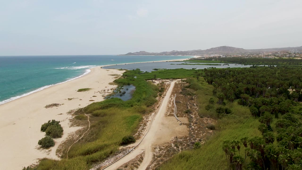 Aerial view at Puerto Los Cabos Beach with the dirt road more centered, lagoon water beside the sand, ocean surf on the left, green palms on the right, and no visible people.
