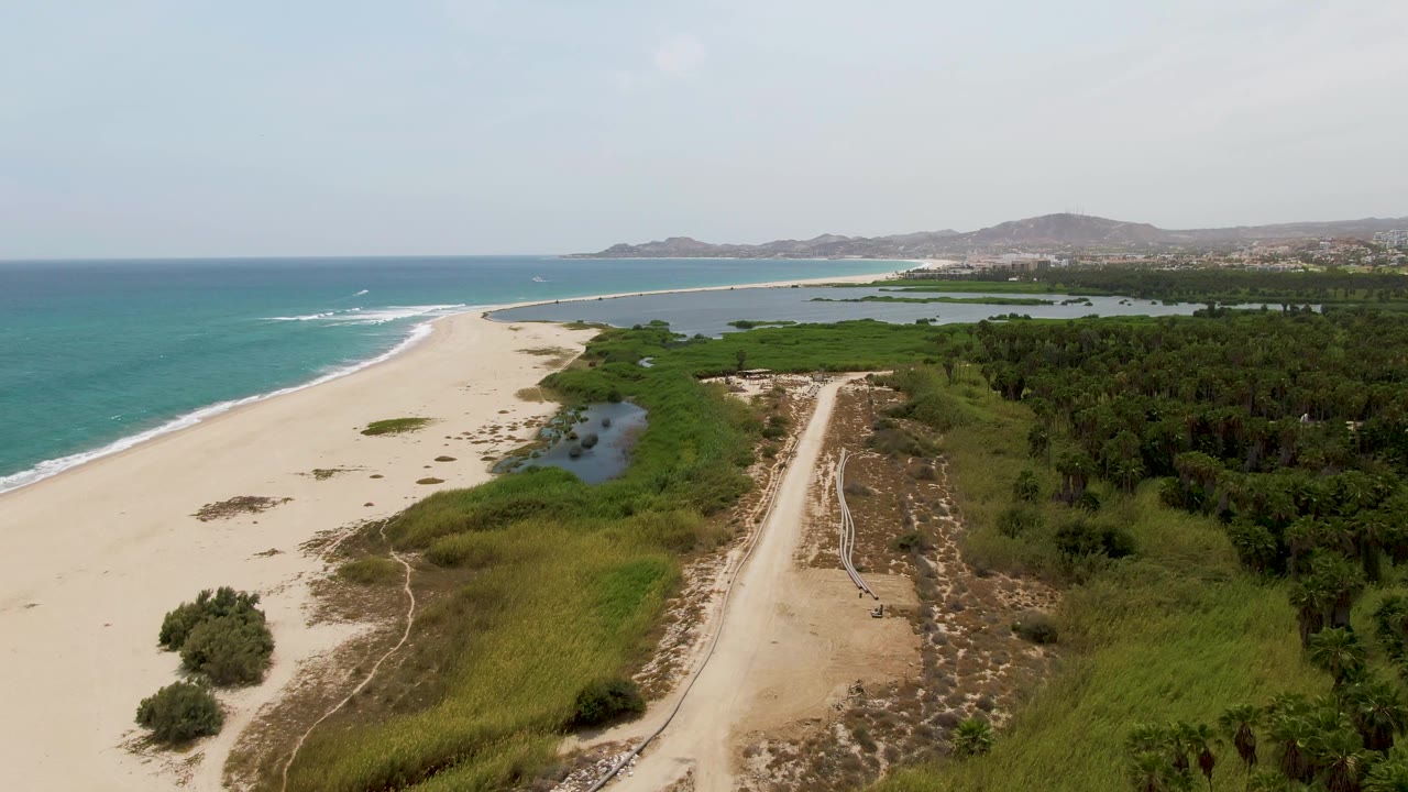 Puerto Los Cabos Beach aerial frame with the beach curving toward distant hills, lagoon water below center, a pale dirt road at right, green vegetation, and no visible people.