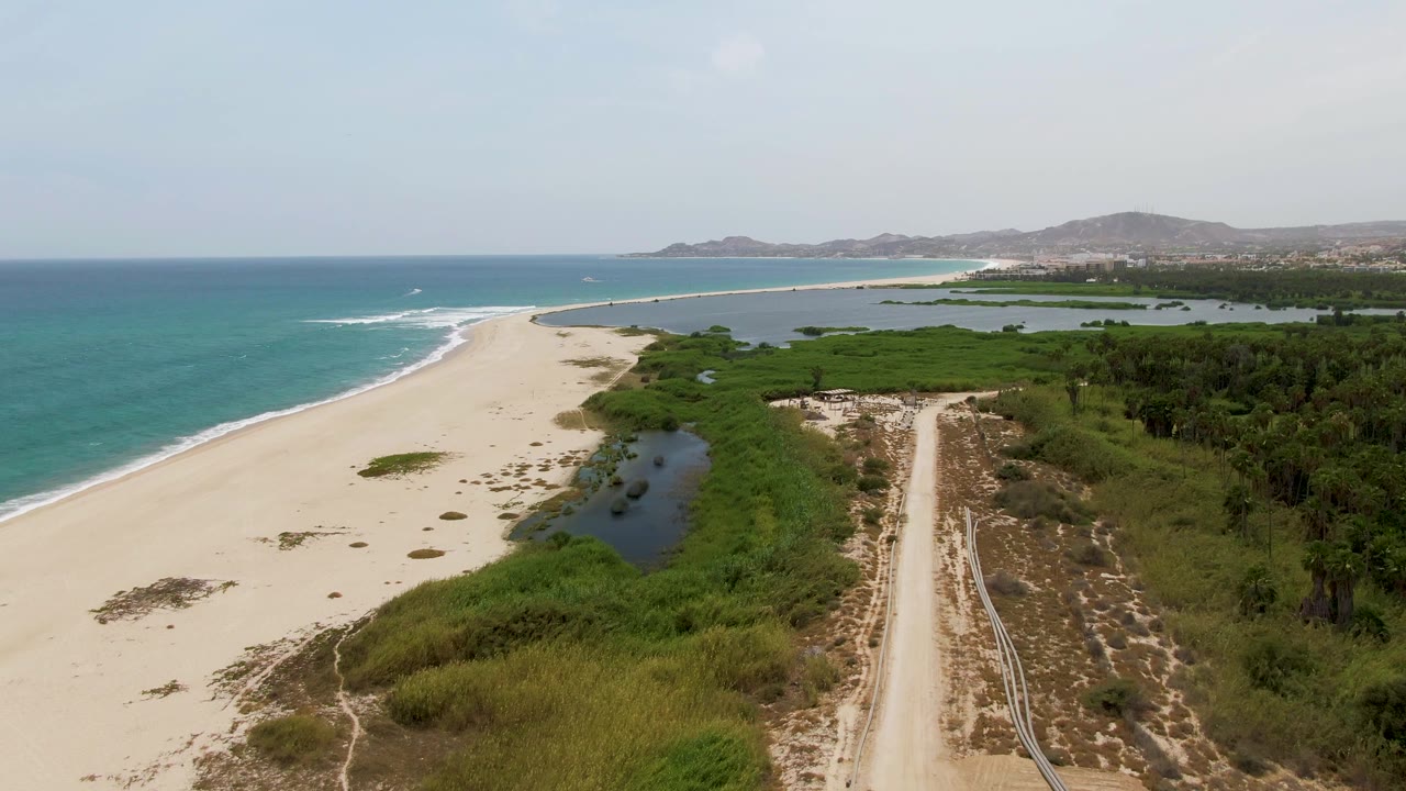 Aerial frame over Puerto Los Cabos Beach with turquoise ocean at left, a broad sandy shore, green wetlands, a pale dirt road at right, distant hills, and no visible people.