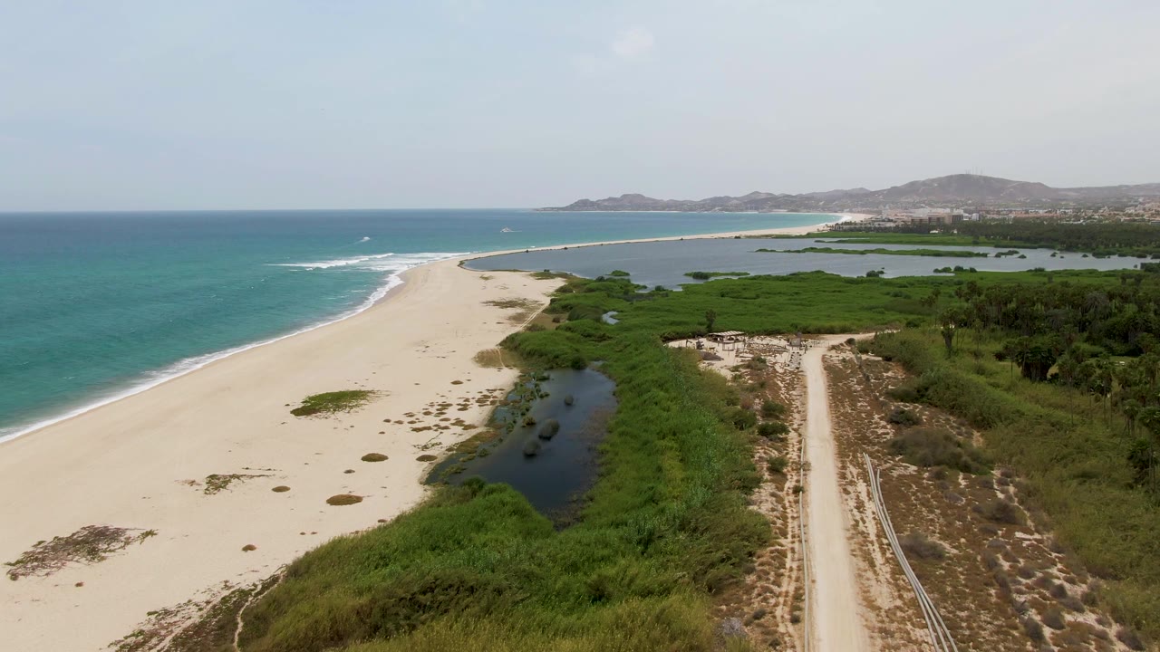 An aerial view over Puerto Los Cabos Beach shows the sandy beach and blue-green ocean on the left, wetland vegetation and water in the center, the dirt road to the right, and no visible people.