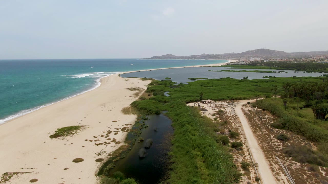 Aerial frame at Puerto Los Cabos Beach showing turquoise ocean on the left, a wide sandy shore, dark lagoon water in the foreground channel, green vegetation to the right, distant hills, and no visible people.