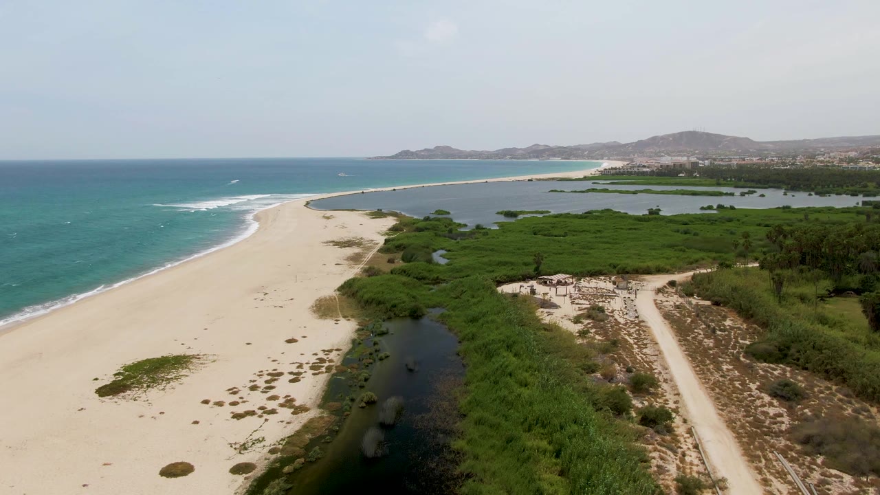 At 0:01, Puerto Los Cabos Beach remains framed from above with the surf edge shifted along the left beach, lagoon pools cutting into the sand and greenery, a dirt access track on the right, and no visible people.