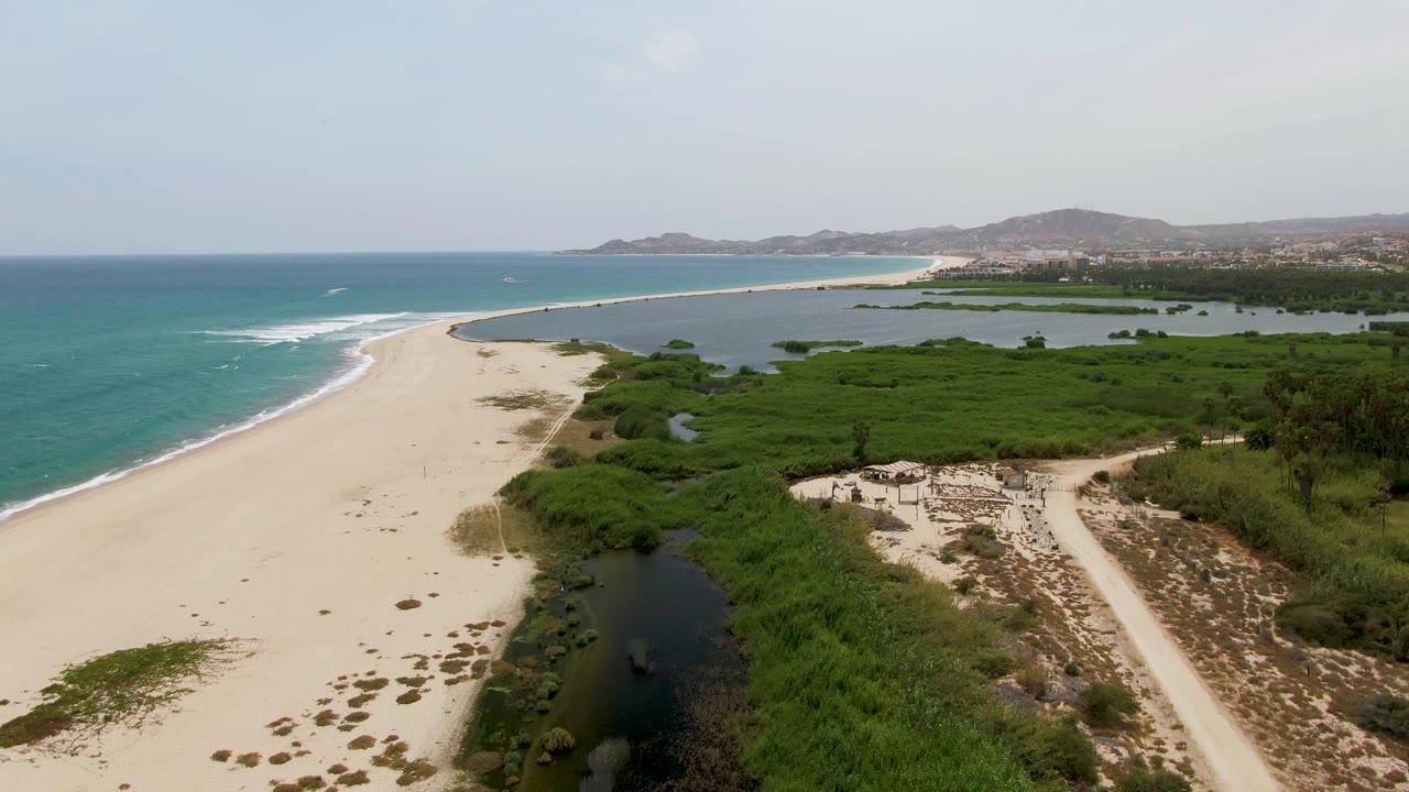 Elevated coastal view of sandy beach, surf, lagoon channel, green shrubs, and distant coastline