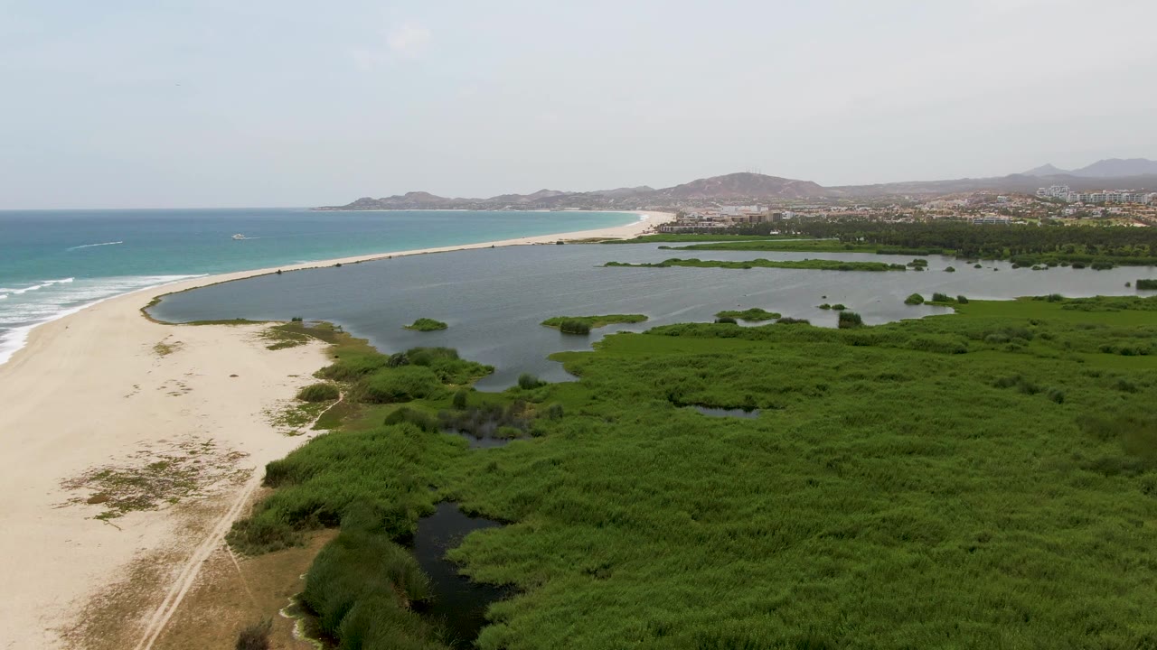 Aerial frame from Puerto Los Cabos Beach with ocean waves lining the left shore, lagoon water wrapping the sandbar, marsh vegetation filling the foreground, distant buildings, and no visible people.