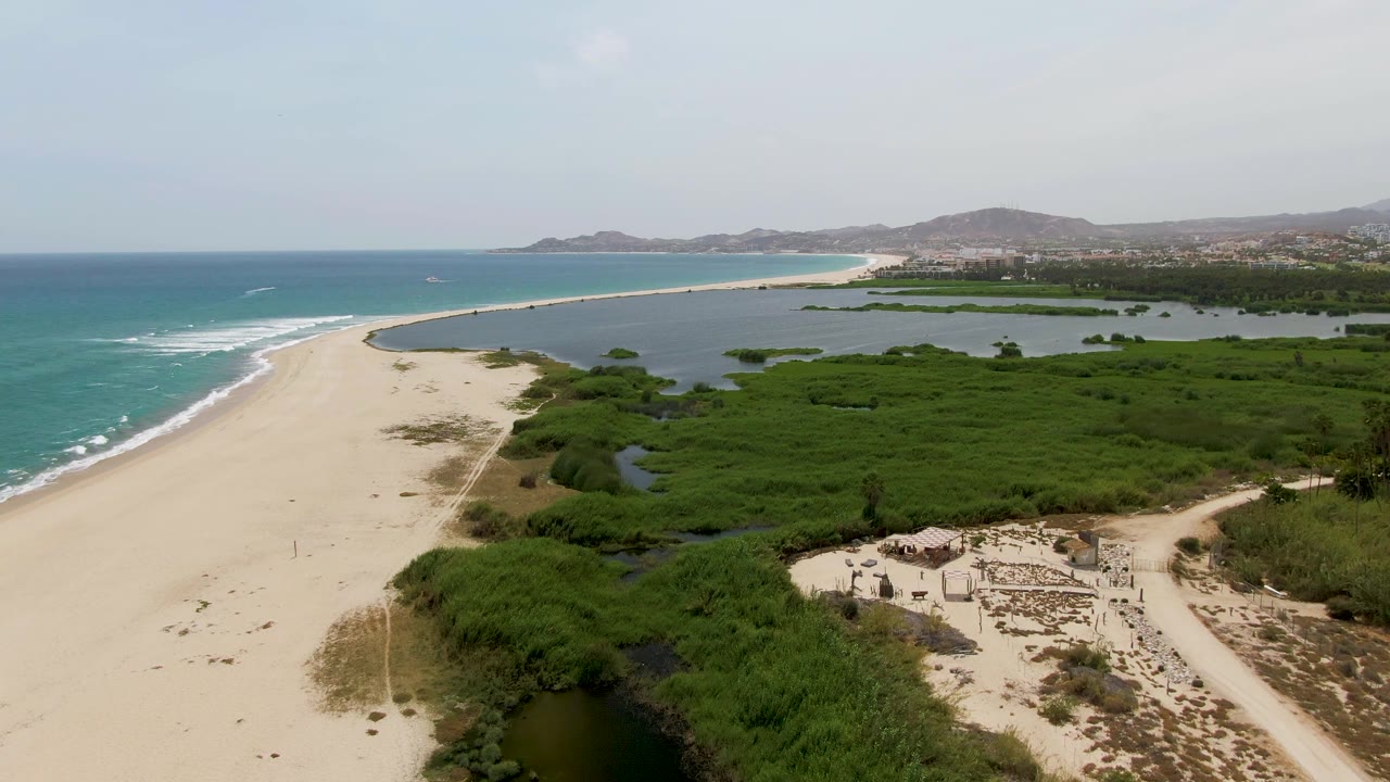 Aerial view at Puerto Los Cabos Beach with the shoreline curving along the ocean, dense green wetland vegetation to the right, small structures near the lower edge, and no visible people.