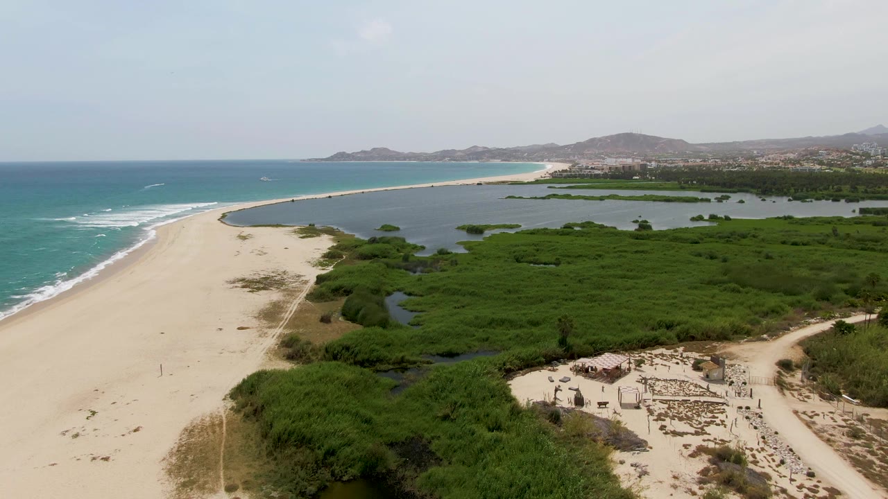 Drone-height frame over Puerto Los Cabos Beach showing a broad sand strip, ocean surf on the left, wetland ponds and greenery on the right, distant hills, and no visible people.