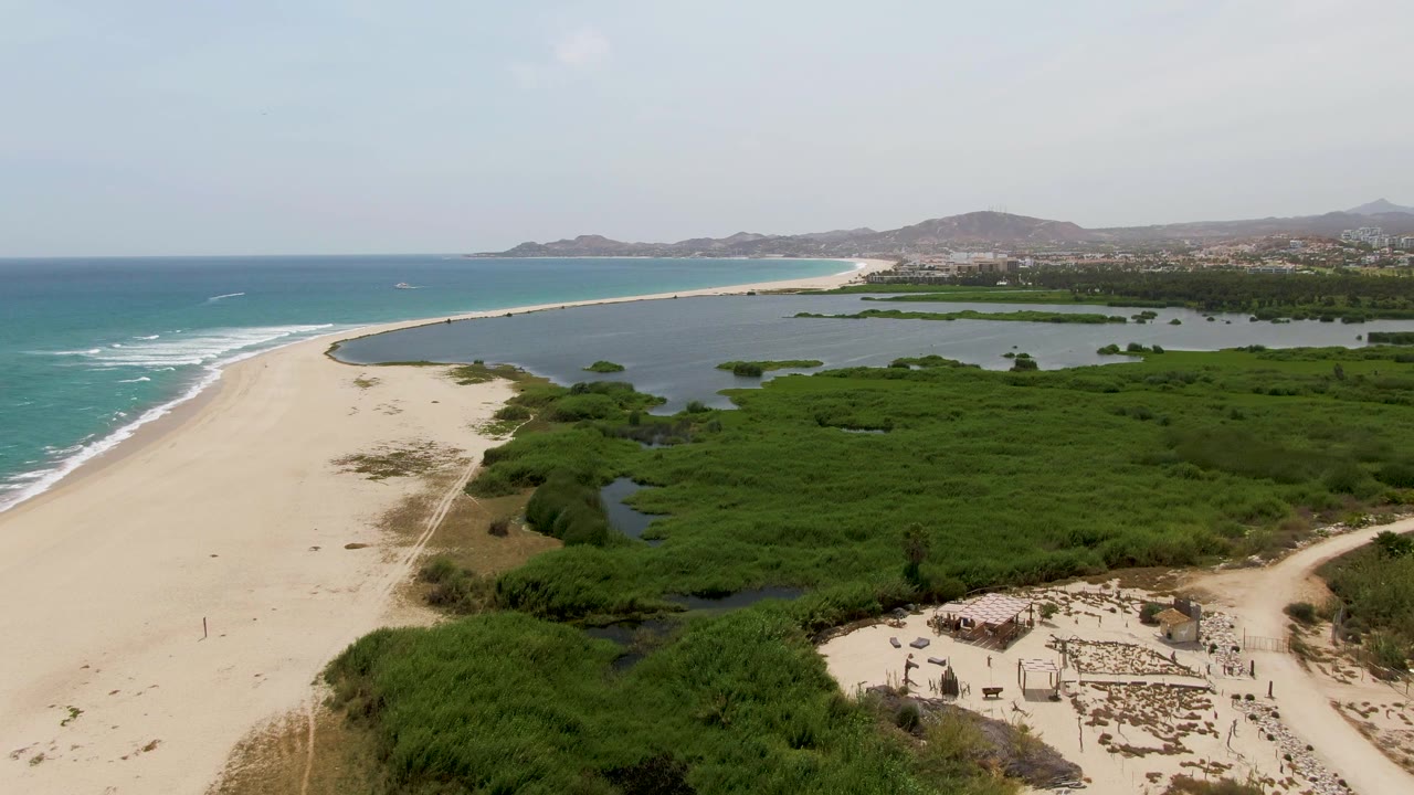 Puerto Los Cabos Beach seen from above with wave lines reaching the sand, a green wetland band beside the beach, scattered small structures near the lower right, and no visible people.