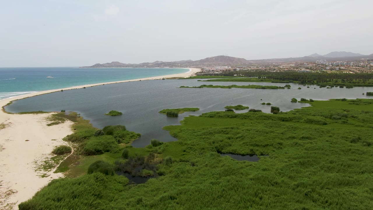 Aerial view at Puerto Los Cabos Beach with a curved sandy edge around lagoon water, thick green marsh in the foreground, blue ocean beyond, distant mountains, and no visible people.