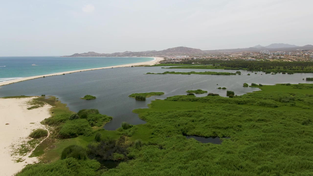 Puerto Los Cabos Beach aerial frame showing lagoon pools within dense green vegetation, a pale sand strip, turquoise ocean to the left, low mountains on the horizon, and no visible people.