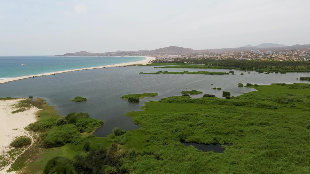 Aerial frame over Puerto Los Cabos Beach with more open lagoon water across the center, marsh vegetation below, a long sandy beach beside the ocean, distant coastal development, and no visible people.