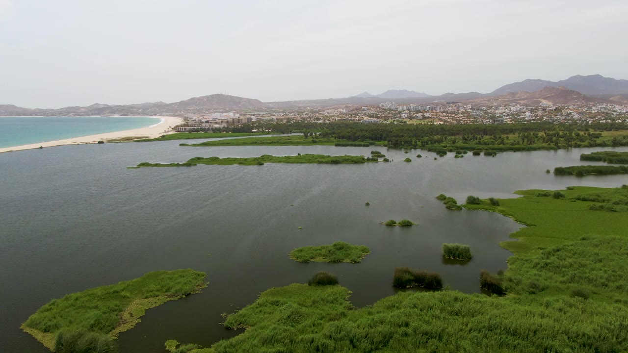 At 0:01, the aerial frame shows broad lagoon water with scattered green marsh patches, Puerto Los Cabos Beach curving at left, town buildings beyond, mountains, and no visible people.