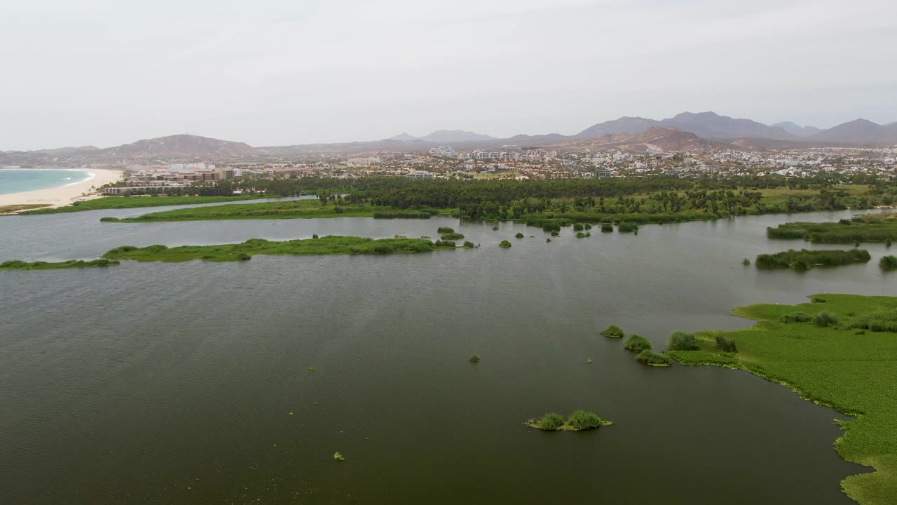 A second Puerto Los Cabos Beach aerial frame looks across broad dark water and scattered green marsh patches toward the pale beach, distant town, hazy mountains, and no visible people.