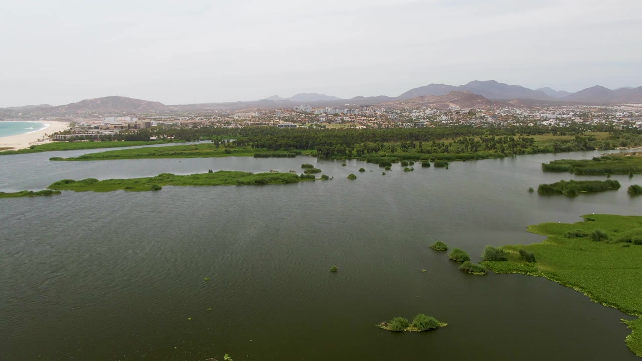 Mid-clip aerial view at Puerto Los Cabos Beach with green marsh islands breaking up calm water, a sandy shoreline on the left, town buildings in the distance, mountains beyond, and no visible people.