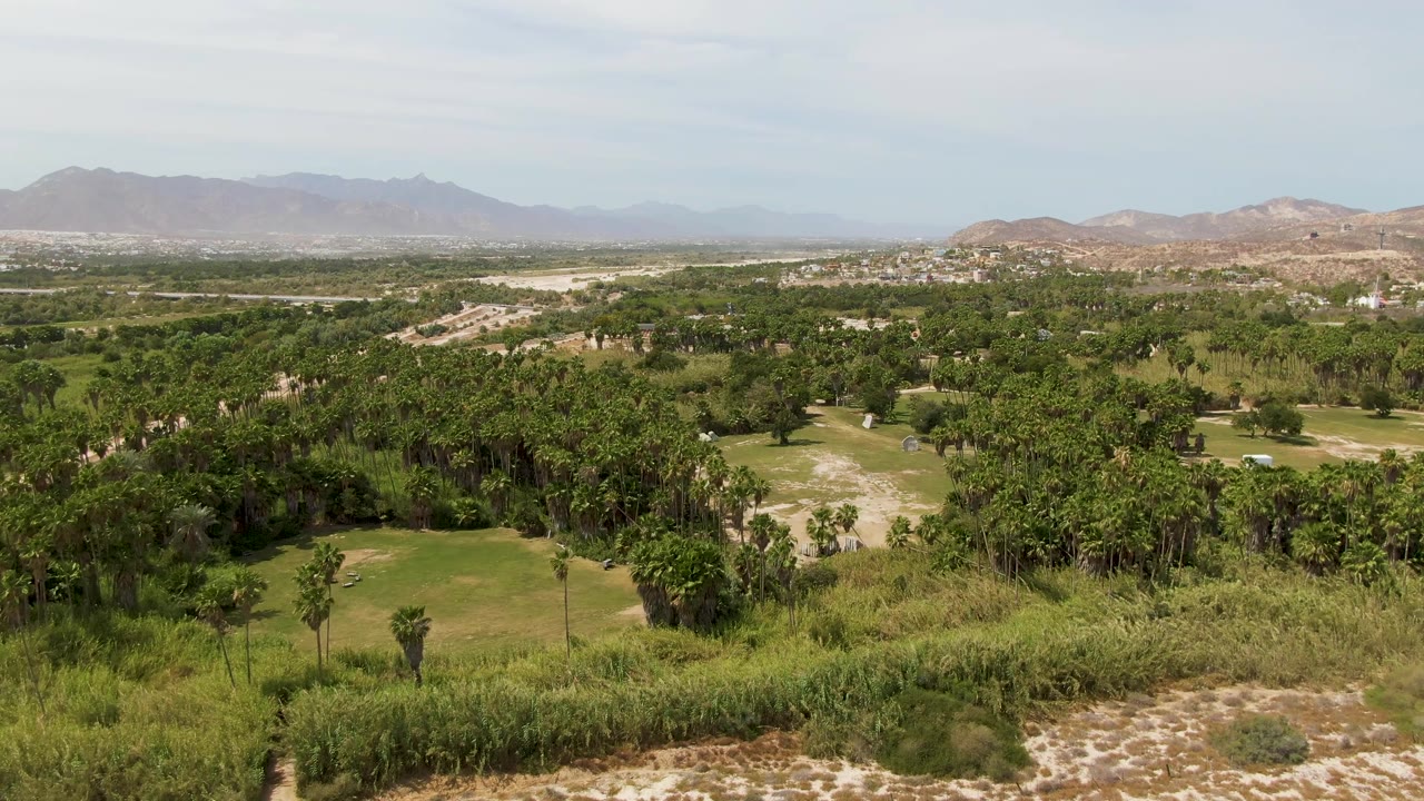 Aerial frame from Puerto Los Cabos Beach, Baja California Sur, with sandy paths in the foreground, green valley vegetation, distant hills and mountains, and no visible people.
