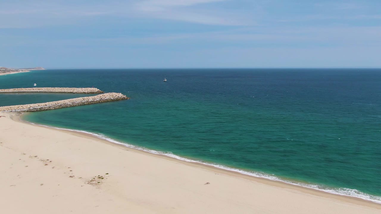 At 0:01, Puerto Los Cabos Beach remains framed from above with the foam line slightly farther along the sand, the breakwater to the left, and no visible people.