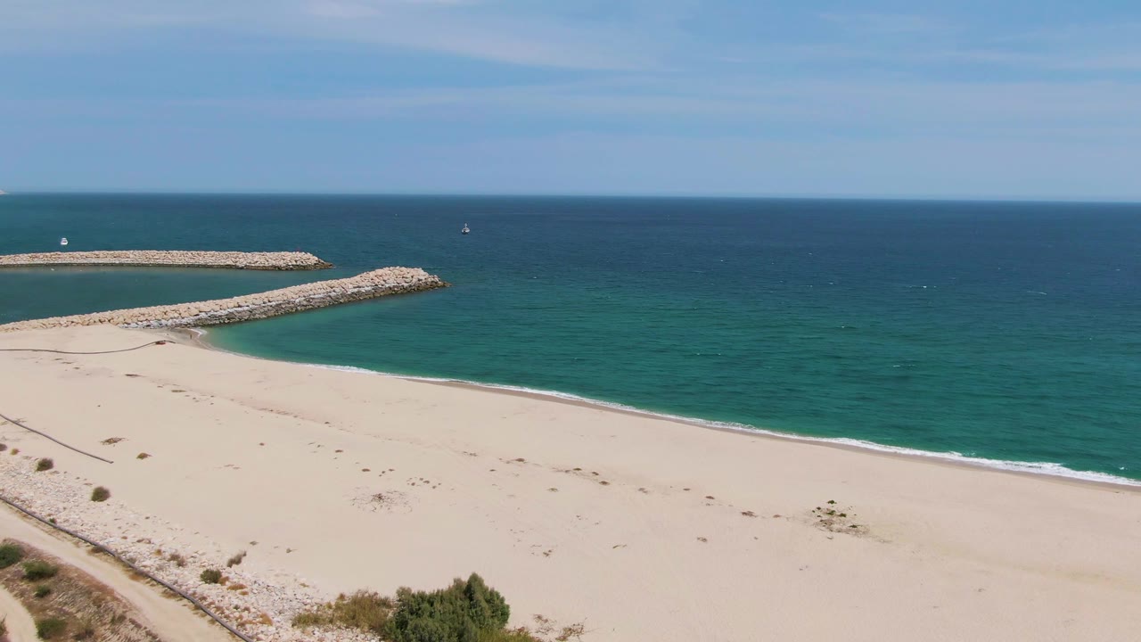 Puerto Los Cabos Beach frame showing a wide sweep of empty sand, a curved rocky breakwater, deeper blue water at the horizon, and no visible people.