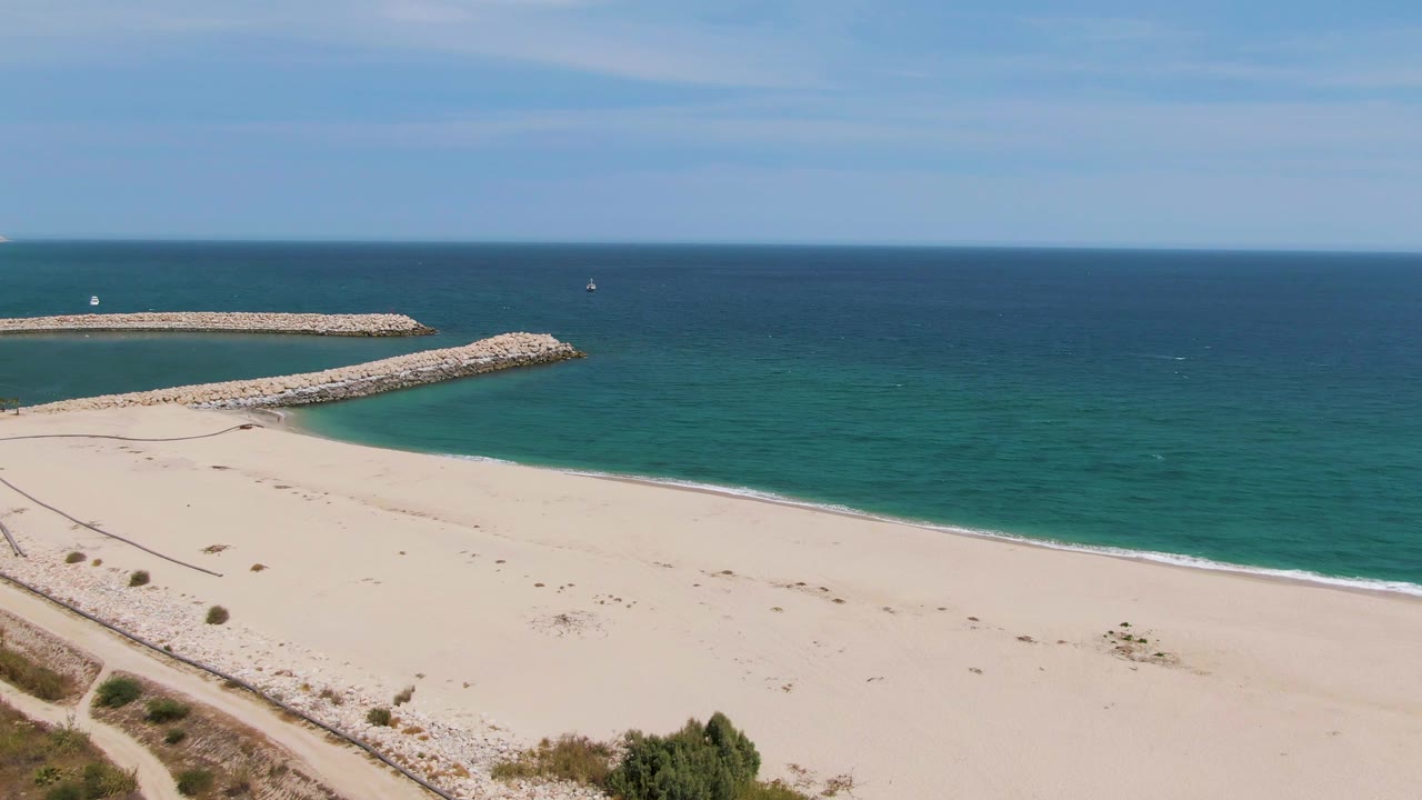 Puerto Los Cabos Beach aerial still with the shoreline running across the lower frame, a rocky jetty extending into turquoise water, and no visible people.