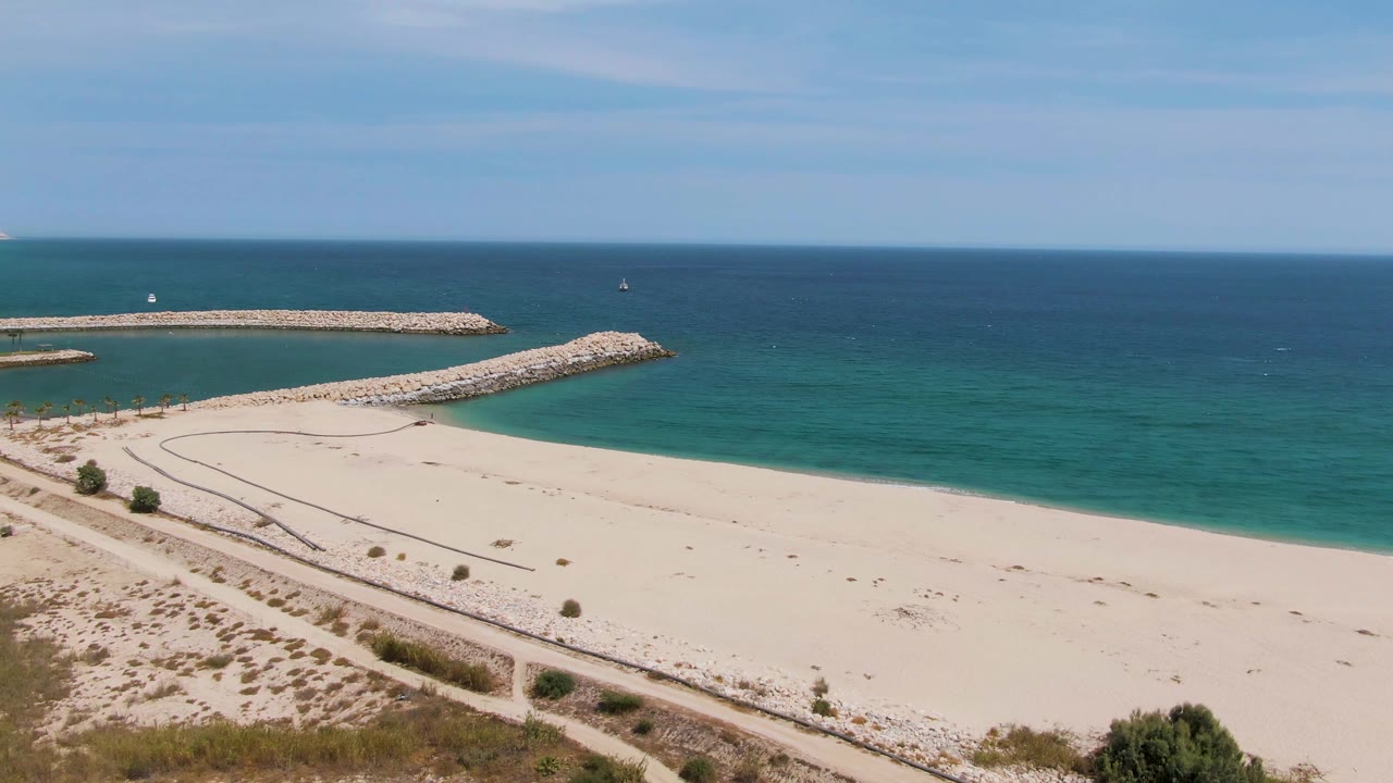Puerto Los Cabos Beach aerial frame with pale sand, turquoise ocean, two rocky breakwaters, dune scrub in the foreground, and very small distant figures near the path.
