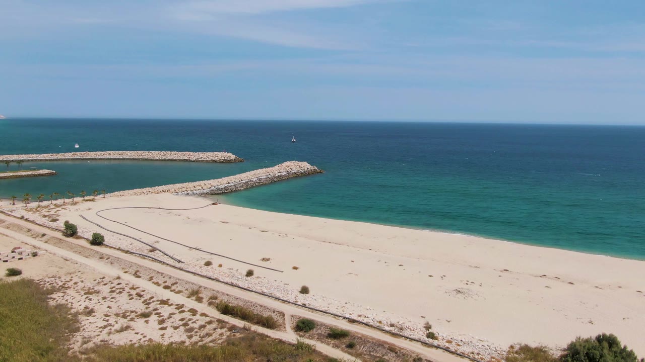 Aerial beach view with rock breakwaters, calm ocean, dune path, and sparse greenery.