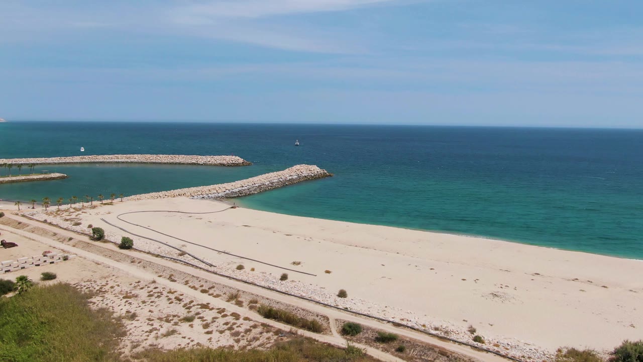 Puerto Los Cabos Beach frame with more dune greenery visible at lower left, small distant people along the path area, open sand, and the breakwaters enclosing calm turquoise water.