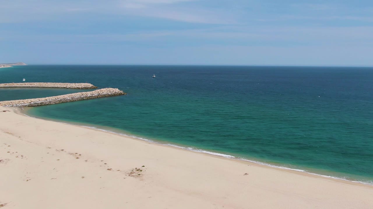 Sandy beach, turquoise ocean, and rocky breakwater under a partly cloudy sky.