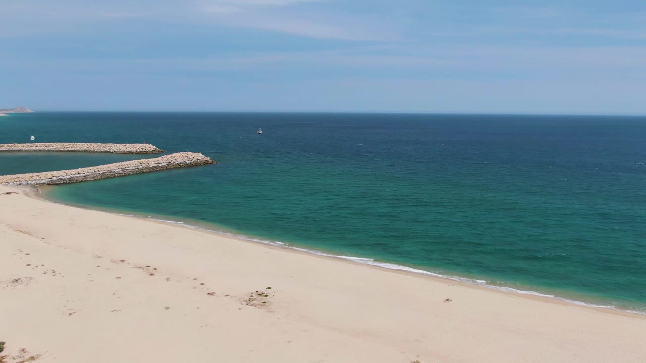 Puerto Los Cabos Beach frame showing a broad empty sand foreground, curved rocky breakwater, blue-green ocean, faint boats offshore, and no visible people.