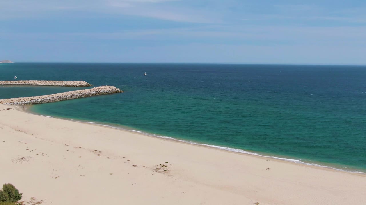 Puerto Los Cabos Beach view with the surf line running along the sand, the rocky breakwater near the upper left, open ocean beyond, and no visible people.