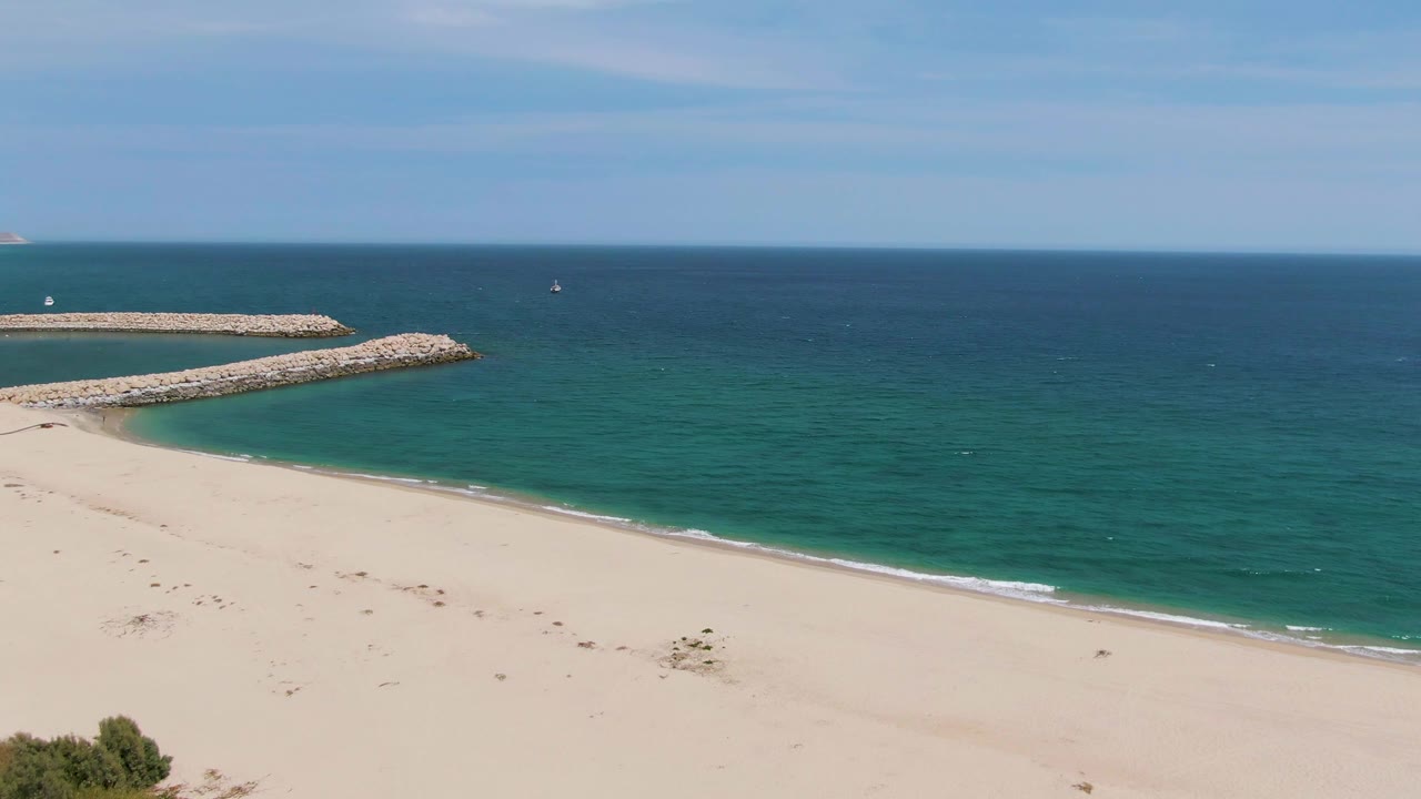 Puerto Los Cabos Beach frame where a small foreground bush enters along the lower edge, with sand, turquoise water, rocky breakwater, distant boats, and no visible people.