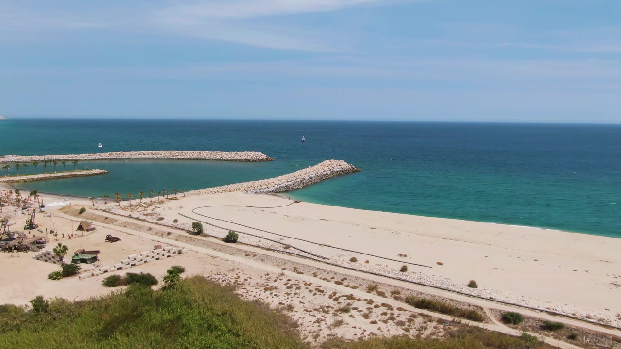 Puerto Los Cabos Beach aerial frame with foreground greenery, broad pale sand, rocky breakwaters, turquoise shallows, deep blue ocean, and tiny beachgoers near umbrellas on the left.