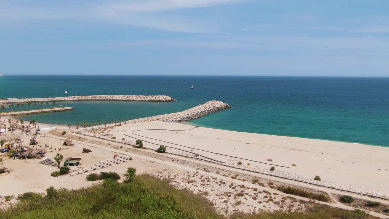 Aerial beach view with rocky jetties, calm turquoise water, umbrellas, palms, and dunes.