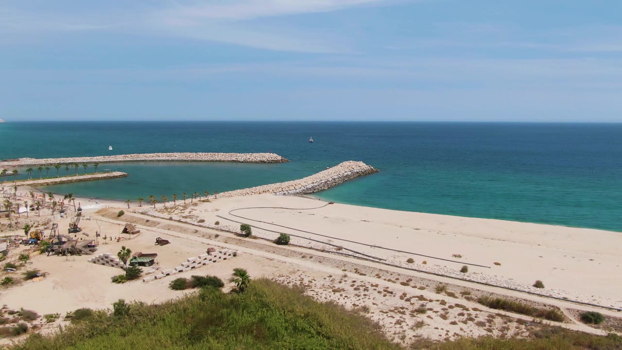 Puerto Los Cabos Beach frame with the breakwater arms spanning the upper beach, calm turquoise water inside the protected area, open sand at right, and small people near umbrellas at left.